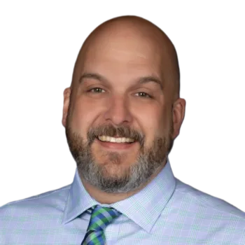 Smiling bald man with a beard, wearing a light blue plaid shirt and green tie, against a dark gradient background.